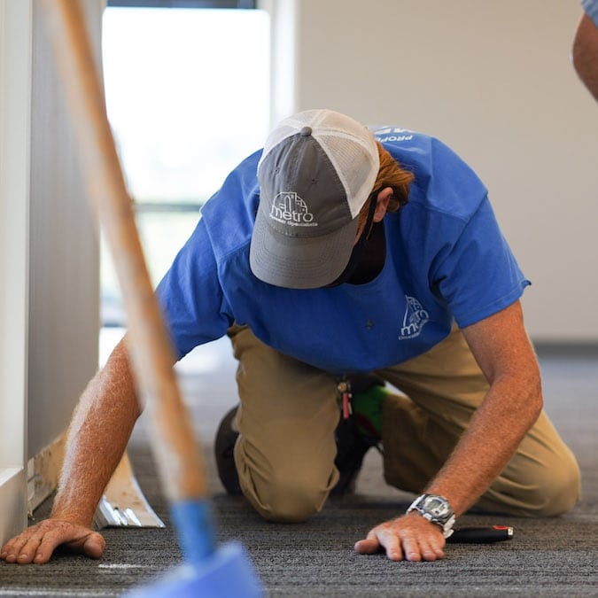 A man in a blue t-shirt kneels on a carpeted floor, checking for water damage.