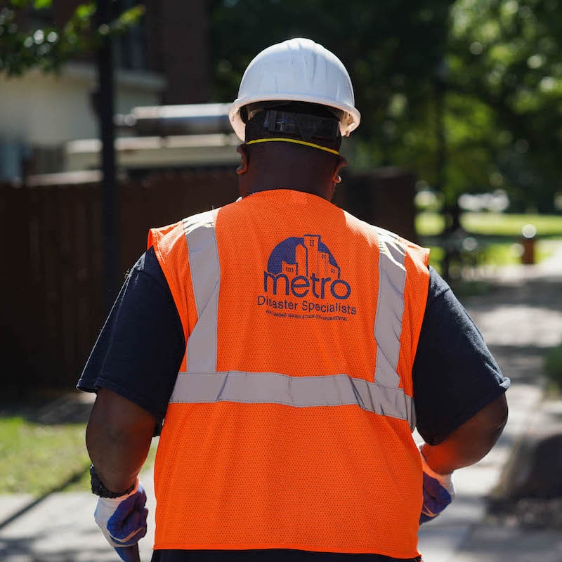A man is walking down a sidewalk, facing away from the camera. He's wearing a white hard hat, a black t-shirt, and an orange workman's vest over the t-shirt. The Metro Disaster Specialists logo is printed on the back of the vest.